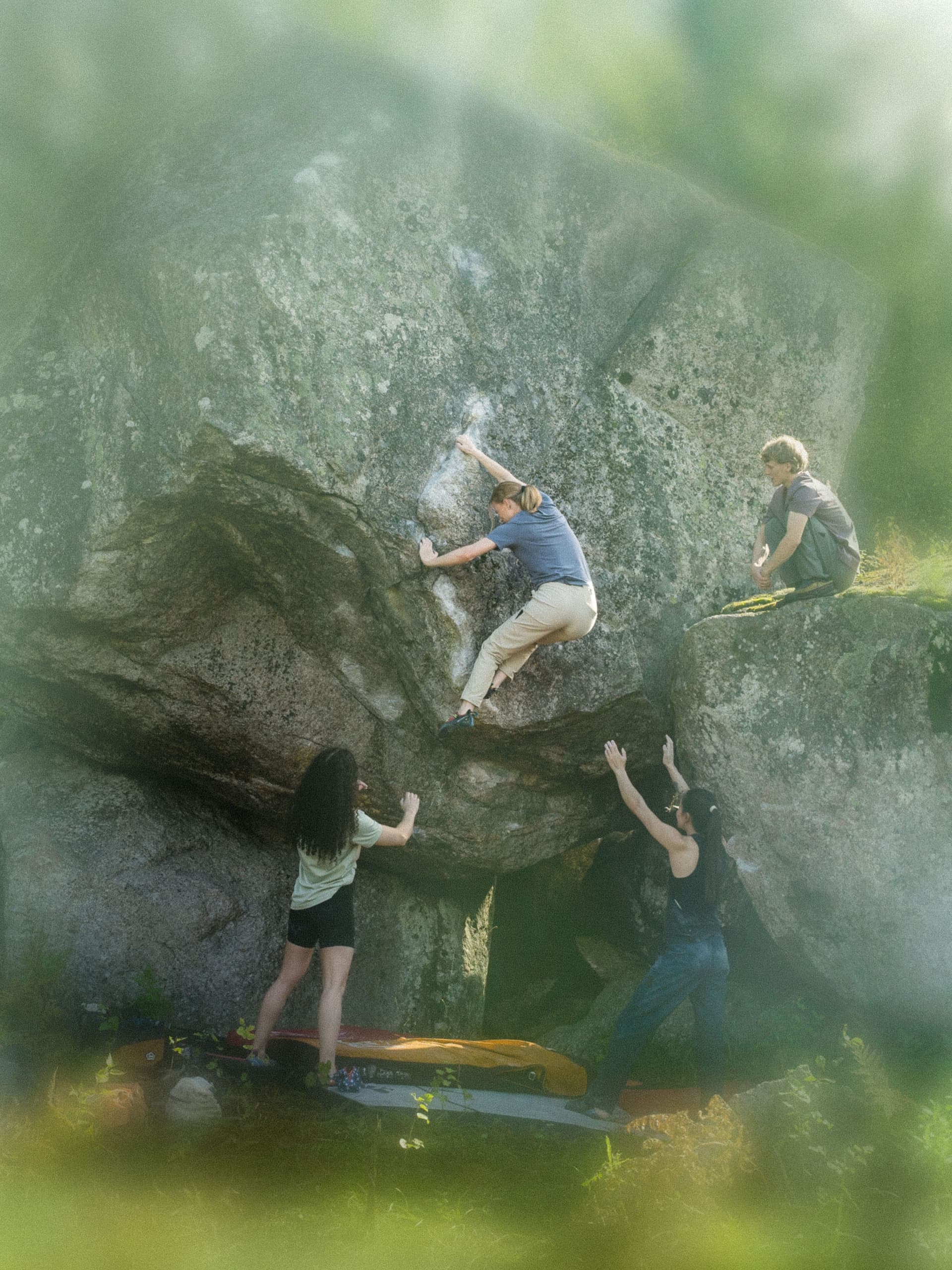 A person boulders on a large rock as three others spot or watch.