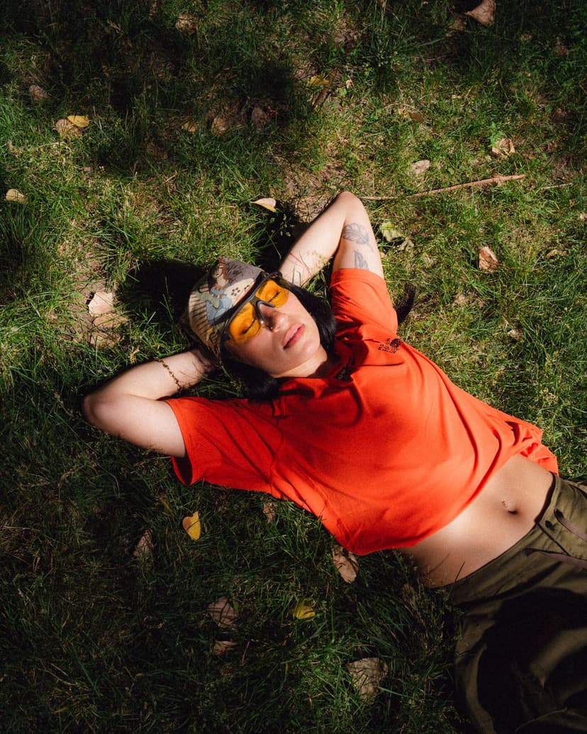 A woman in a patterned hat and yellow sunglasses lies on sun-dappled grass with hands behind her head, wearing an orange crop top and green pants.