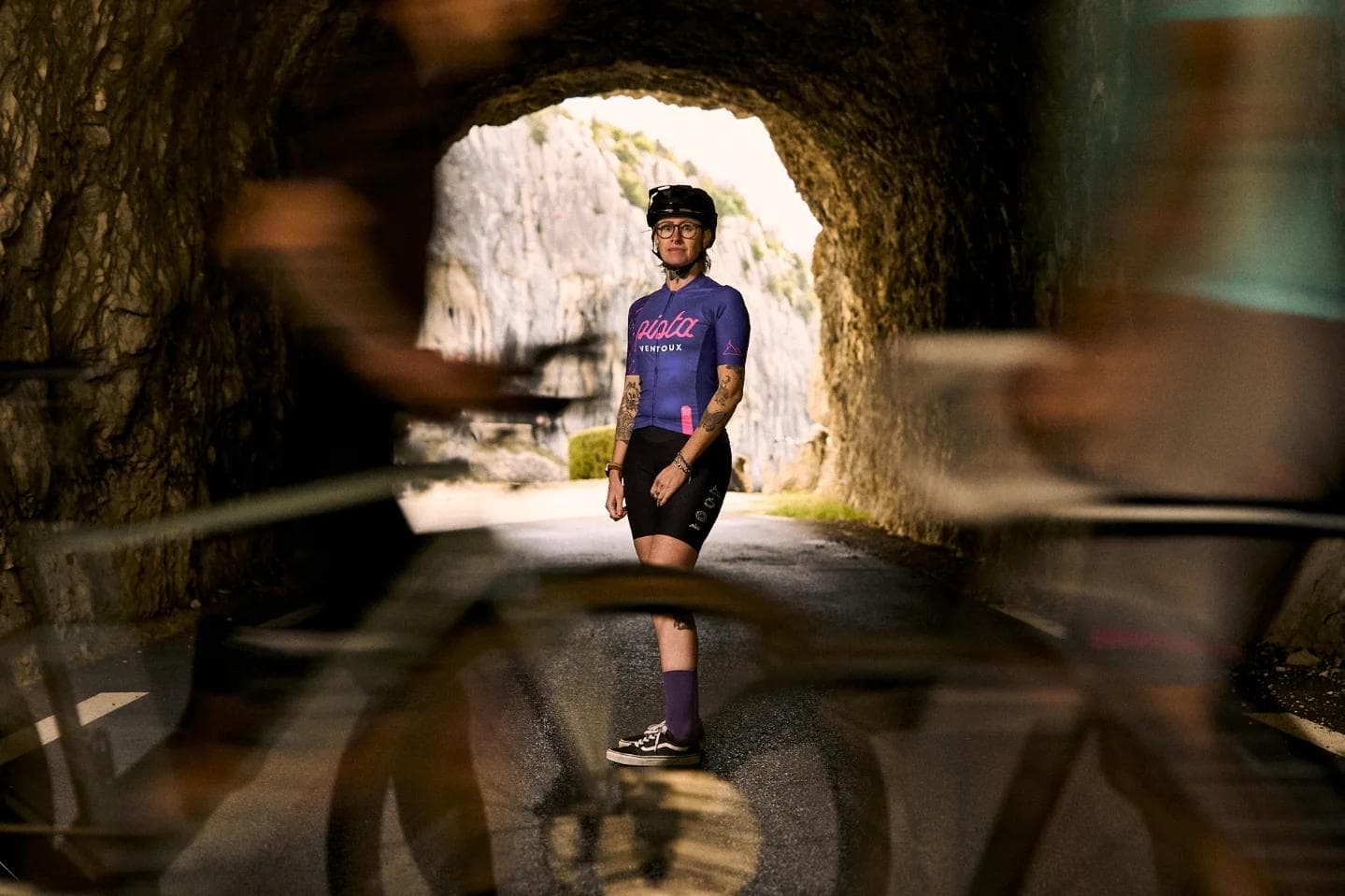 A person in cycling gear stands at a tunnel entrance, looking towards bright cliffs, with blurred cyclists passing in the foreground.