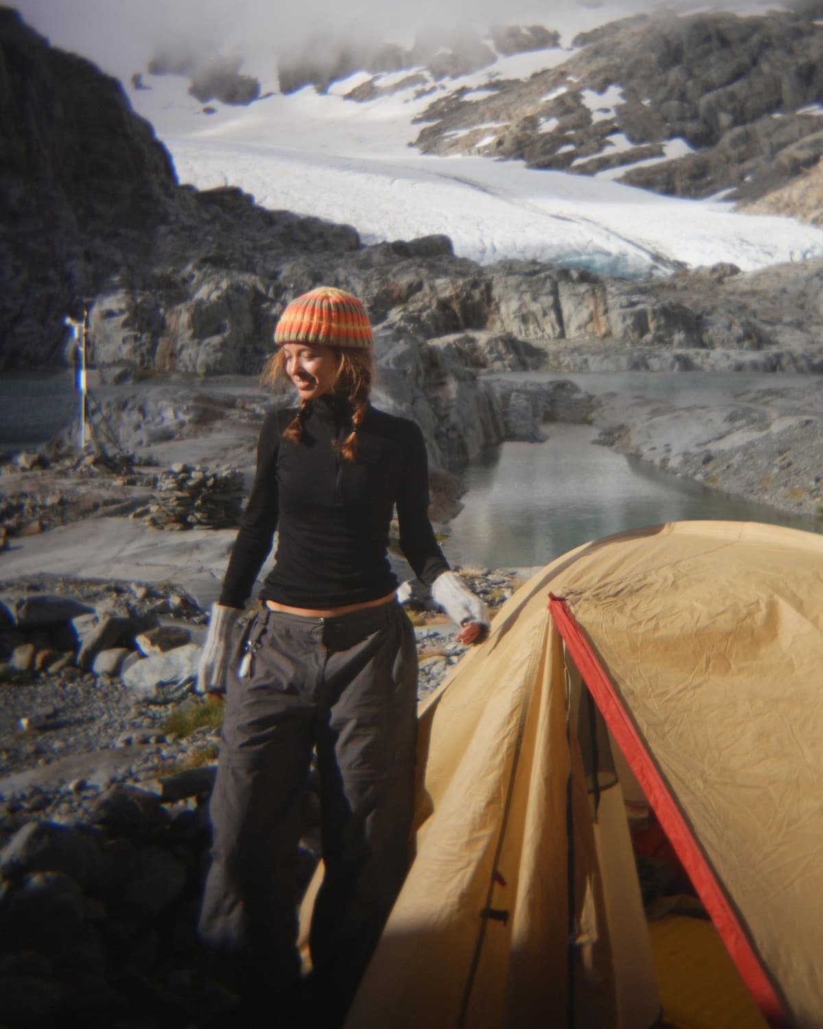 A woman stands next to a tent in a rocky, mountainous glacial landscape with a lake.