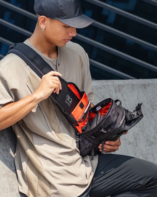 A man wearing a cap and tan shirt puts a Nintendo Switch into a black sling bag with an orange interior.