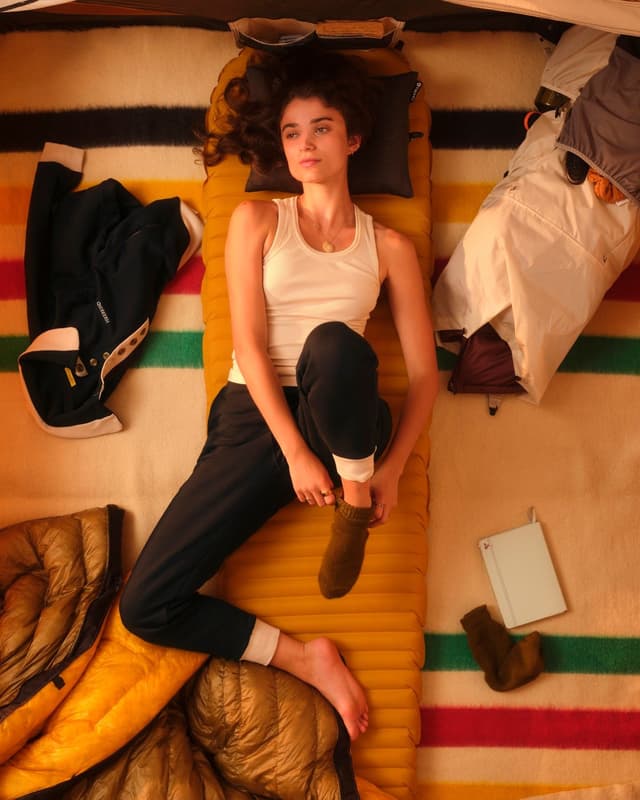 A young woman with curly hair lies on a yellow sleeping pad inside a tent, putting on a green sock, surrounded by camping gear on a striped blanket.