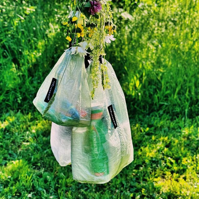 Three translucent bags containing items, hanging from a cluster of wildflowers against a vibrant green outdoor background.