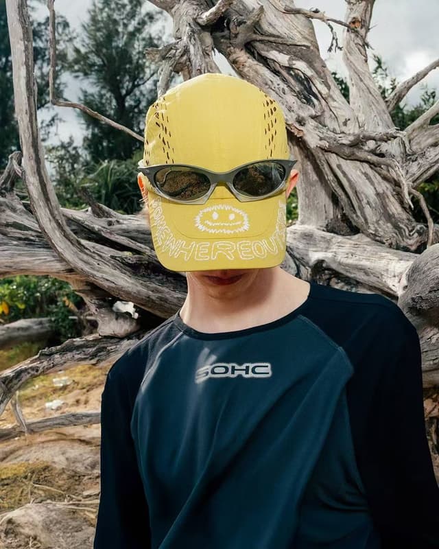 A person wearing a yellow cap with a smiley face and dark sunglasses, and a dark blue long-sleeved shirt, stands before a large, gnarled tree.