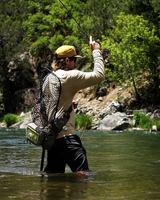A man in a hat and sunglasses stands in a river, casting a fly fishing rod.