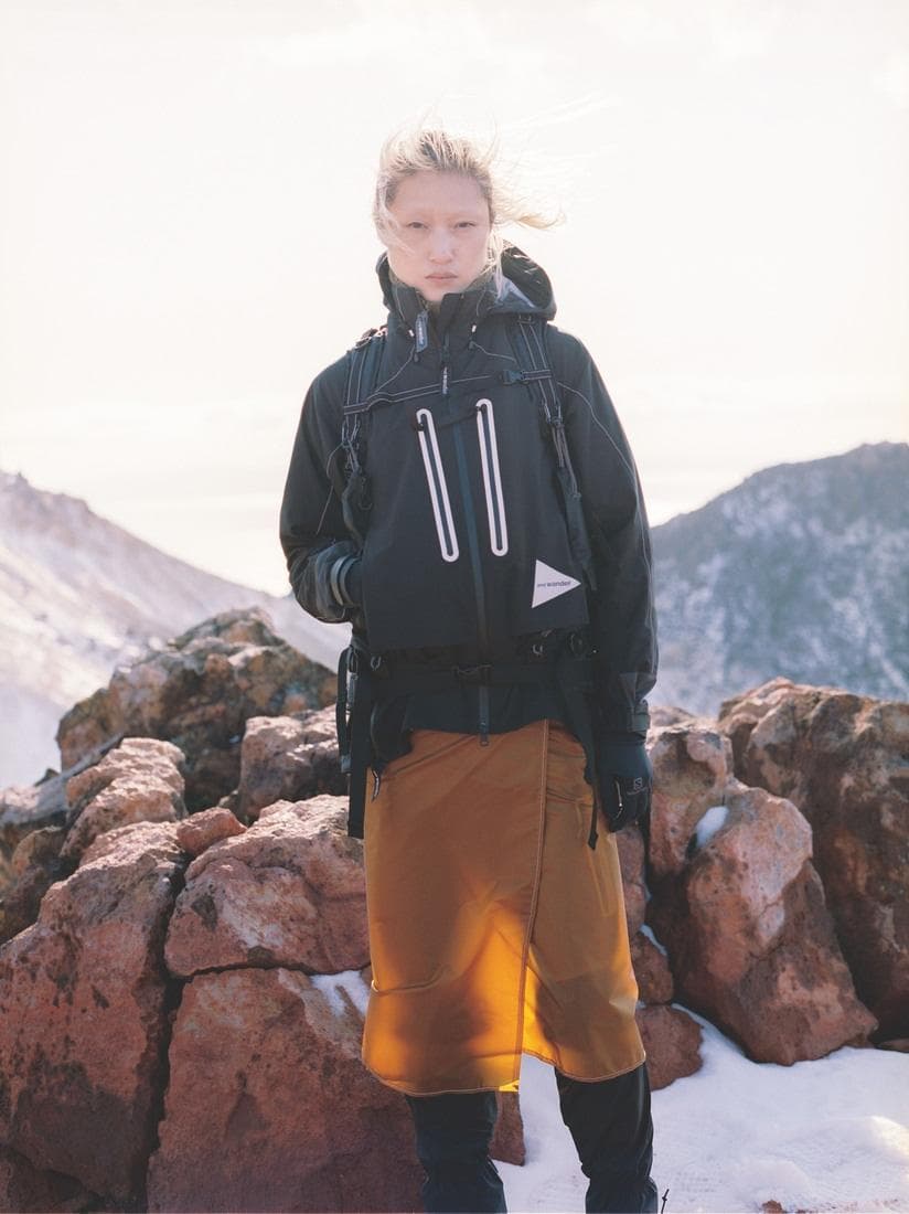 A blonde person in a dark hiking jacket, backpack, and yellow wrap-around skirt stands on a snowy, rocky mountain.