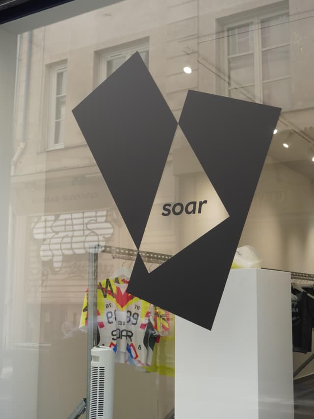 Store window with a dark geometric "soar" logo, colorful athletic apparel, and building reflections.