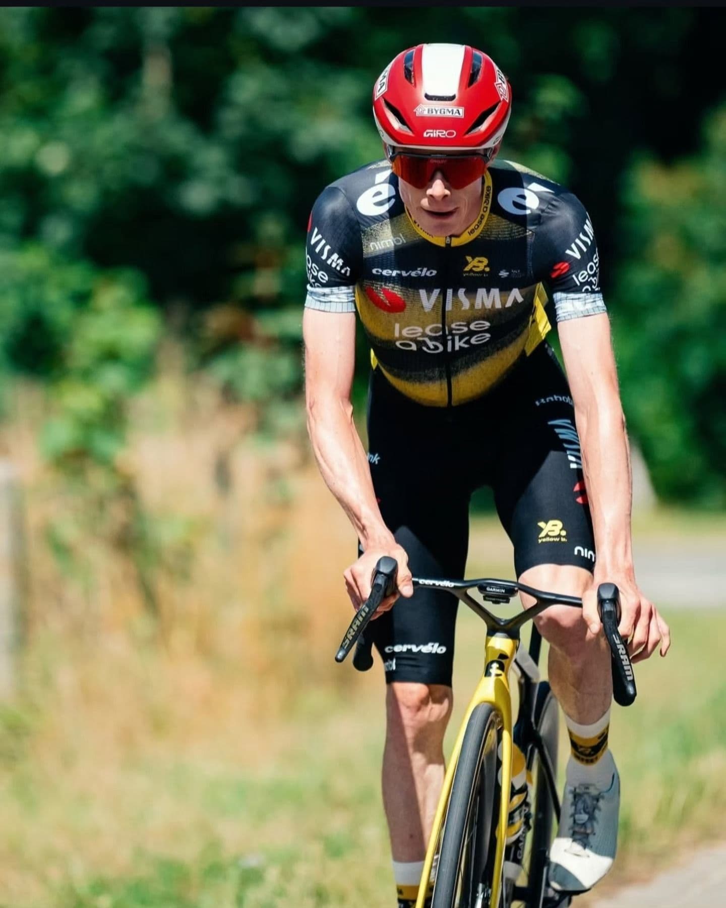 A male cyclist in a black and yellow team kit, red helmet, and sunglasses rides a yellow road bike outdoors.