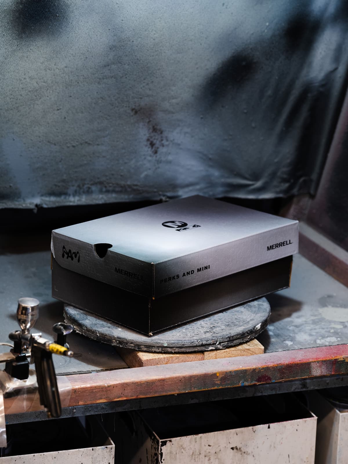 A Merrell Perks and Mini shoe box on a grungy workbench next to an airbrush.