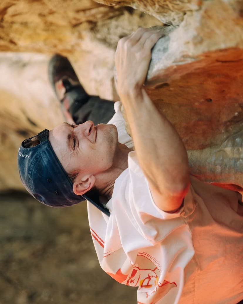 A man in a blue cap and white shirt climbs a rock, gripping a hold with his right hand and looking up.