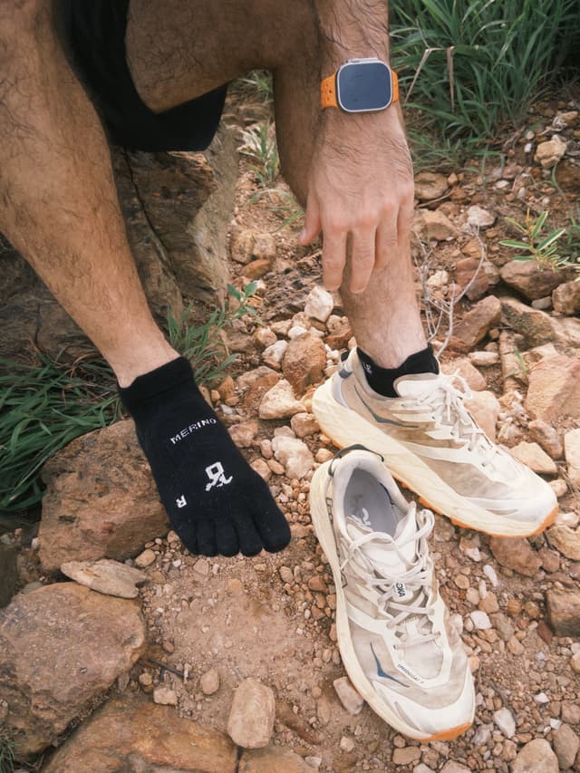 A person's legs and arm show one foot in a black toe sock and two dirty beige trail shoes on rocky ground, with an orange-banded smartwatch on the wrist.
