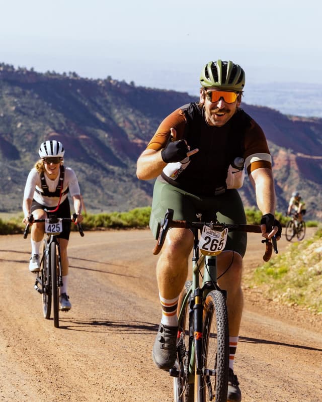 Man on gravel bike smiling and giving a shaka sign on a dirt road, with mountains and other cyclists in the background.