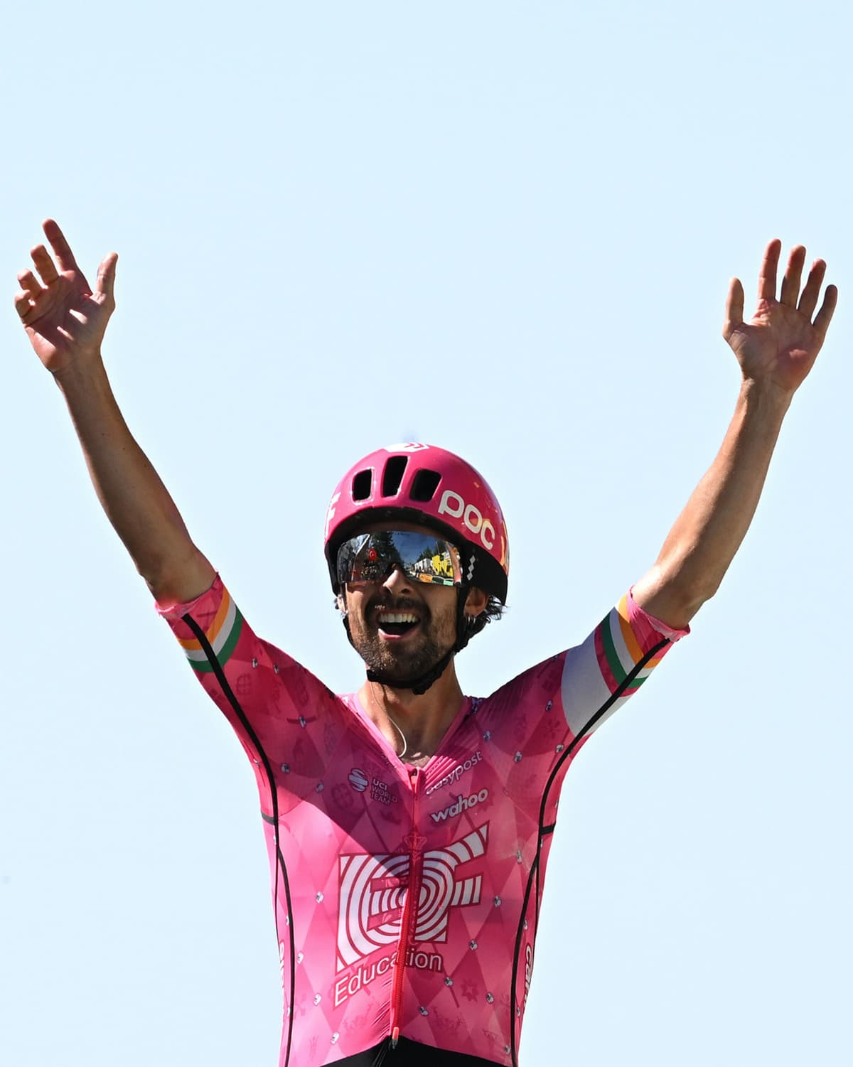 A smiling cyclist in a pink racing kit raises his arms in triumph against a bright blue sky.