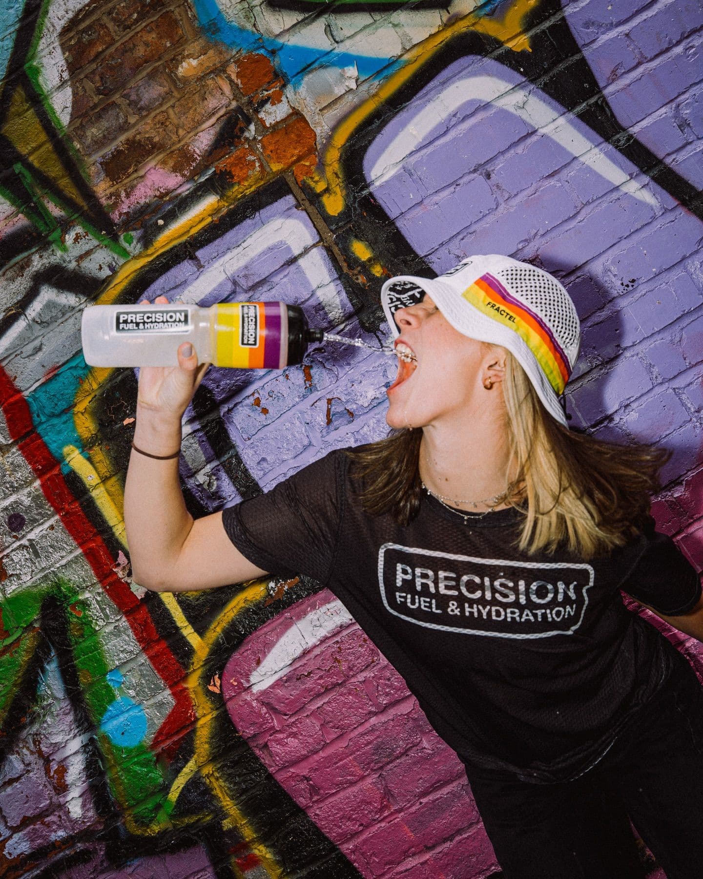 A woman in a Precision Fuel & Hydration shirt and white hat drinks from a branded water bottle, spilling water, against a colorful graffiti wall.