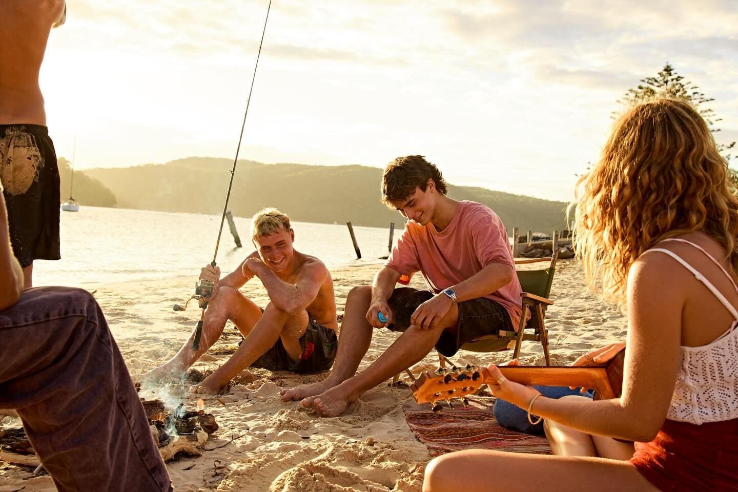 A group of young people gathered around a small campfire on a sandy beach at sunset, with one holding a fishing rod and another playing a guitar.