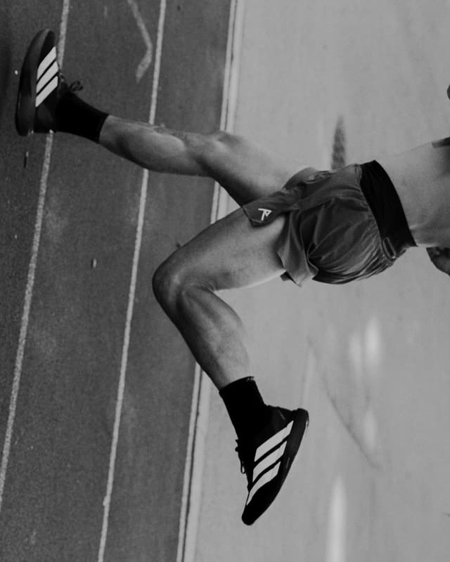 Black and white image of an athlete's legs in stride on a track, wearing shorts and three-striped athletic shoes.