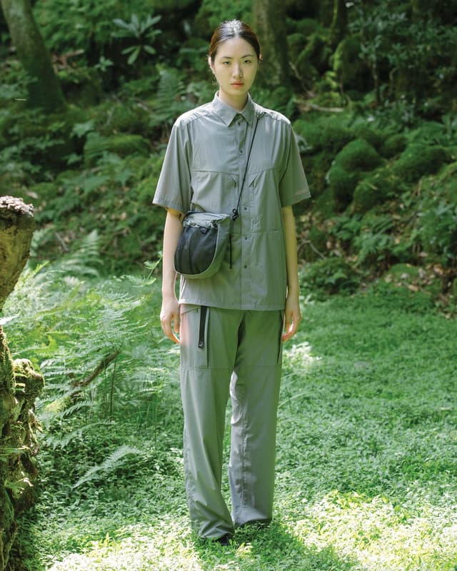 A woman wearing a grey short-sleeve shirt, matching utility pants, and a grey and black crossbody bag stands in a lush green forest.