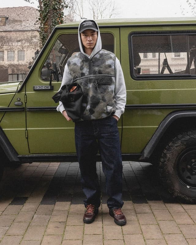 A man in a cap, grey hoodie, and patterned fleece vest stands in front of an olive green G-Wagen, holding a black bag.