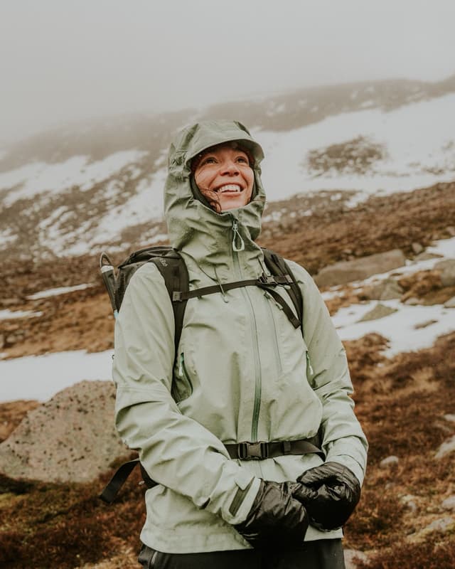 A smiling woman in a green rain jacket, backpack, and gloves looks up in a misty, snowy mountain landscape.