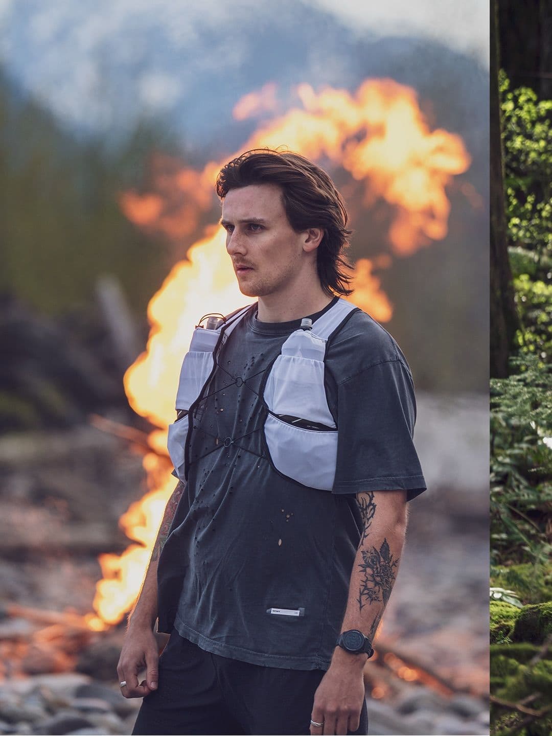 A man in a dark t-shirt and white hydration vest stands outdoors with a large fire on his left and a green forest on his right.