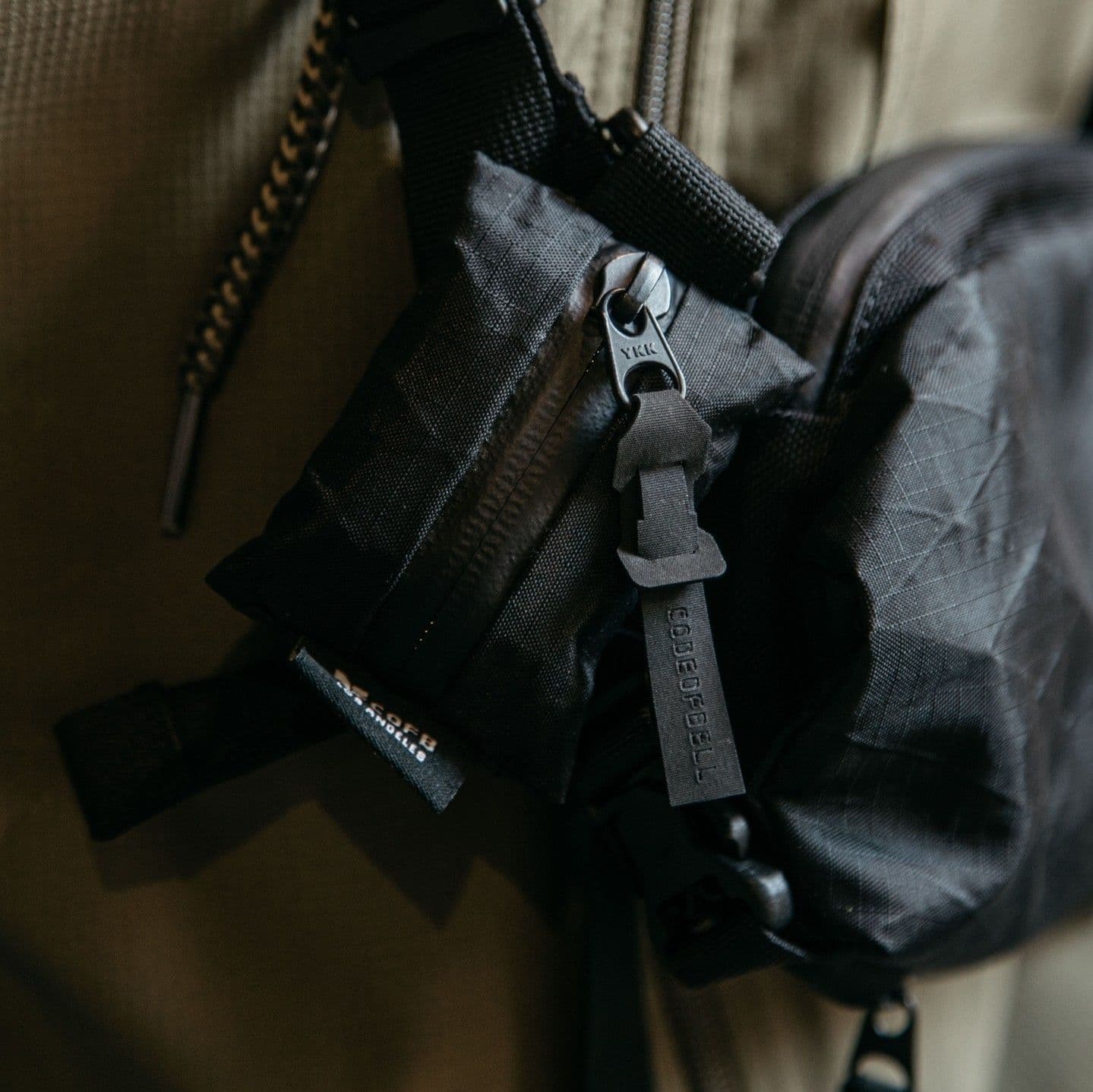 Close-up of a black ripstop bag featuring a YKK zipper and a "CODE OF BELL" branded pull tab.