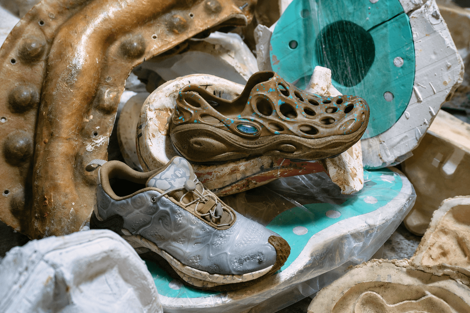 A brown perforated shoe and a grey sneaker lie on a pile of worn molds and industrial materials.