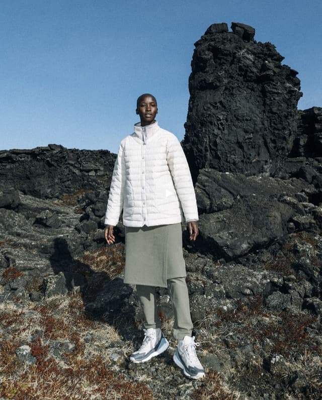A bald person in a white puffer jacket, olive green skirt and pants, and grey hiking shoes stands on rocky terrain under a blue sky.