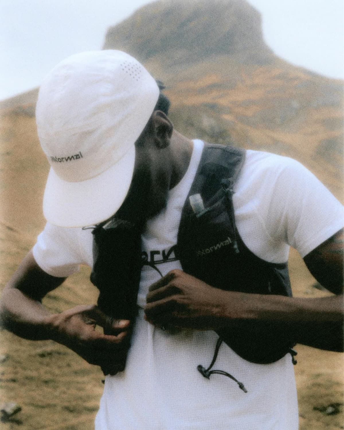 A person in a white "Normal" cap and Norrwal running vest adjusts the vest against a blurry, foggy mountain backdrop.