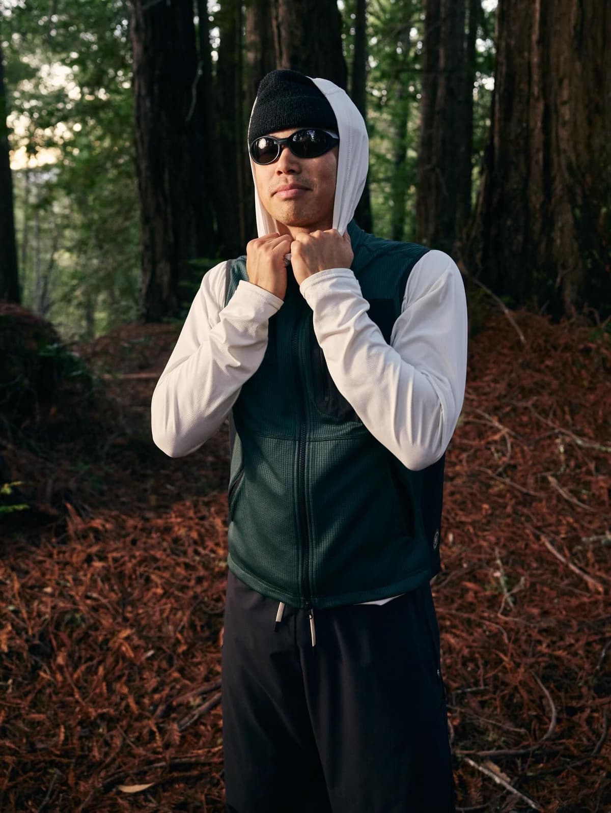 A man in activewear, wearing a black beanie, sunglasses, a white long-sleeve shirt, and a dark green vest, stands in a forest.