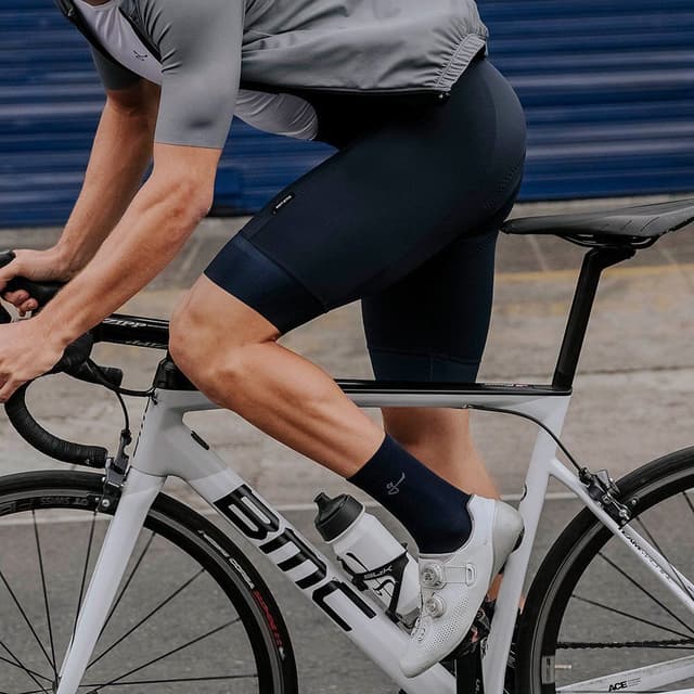 A cyclist in a grey jersey, dark blue bib shorts, and white cycling shoes rides a white BMC road bike.