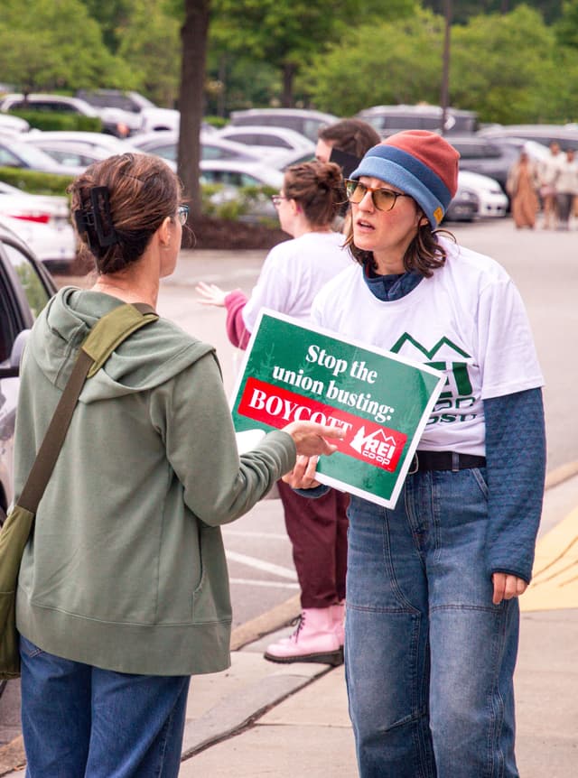 A woman holds a protest sign that reads "Stop the union busting: BOYCOTT REI CO-OP" while speaking to another woman in a parking lot.