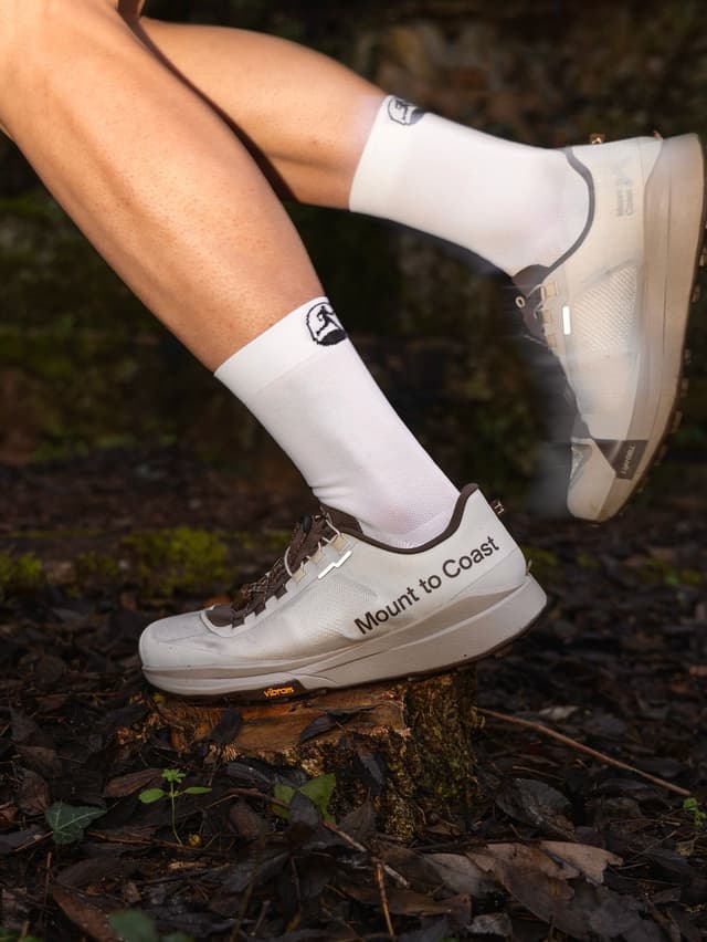 A person's legs wear white socks and light "Mount to Coast" trail shoes, with one foot on a tree stump in a forest.