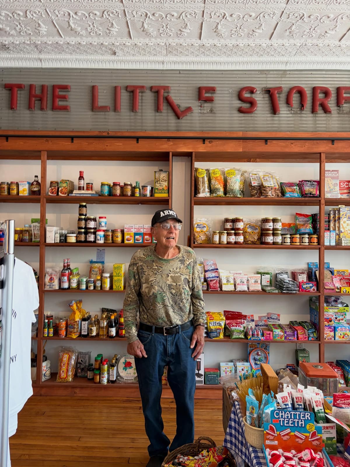 An older man stands in front of shelves packed with goods in "THE LITTLE STORE."