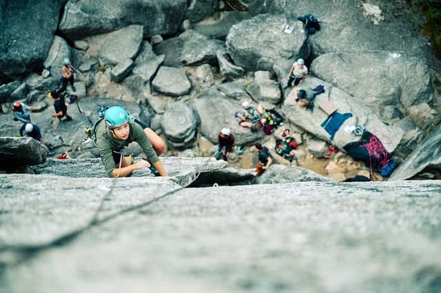 A climber in a teal helmet ascends a rock face, with many other climbers visible on boulders below.