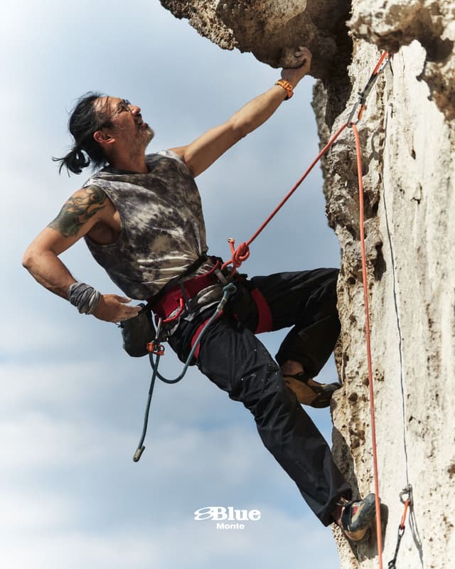 A male climber ascends an overhanging rock face, gripping a handhold with his right hand.
