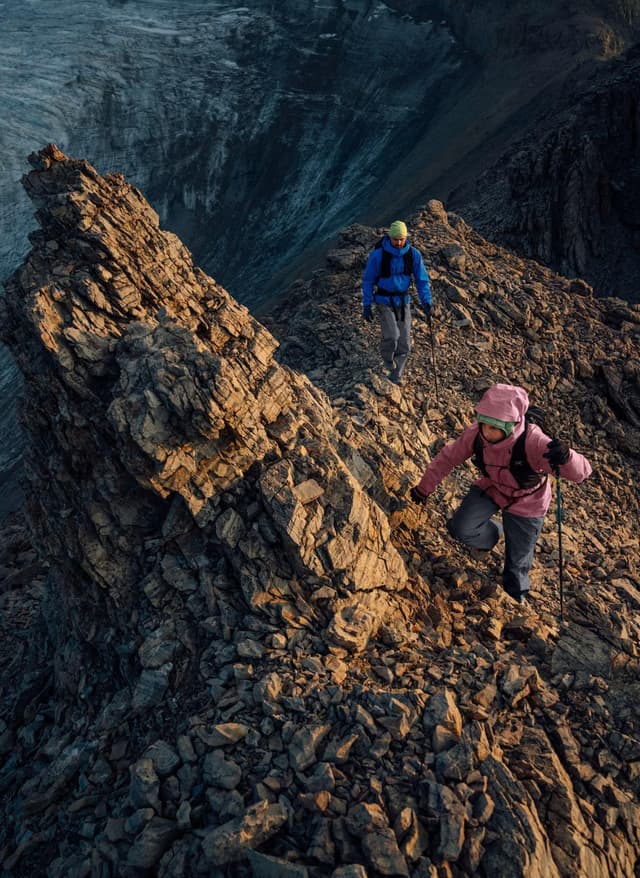 Two hikers ascend a rocky mountain ridge with trekking poles and a glacier in the distance.