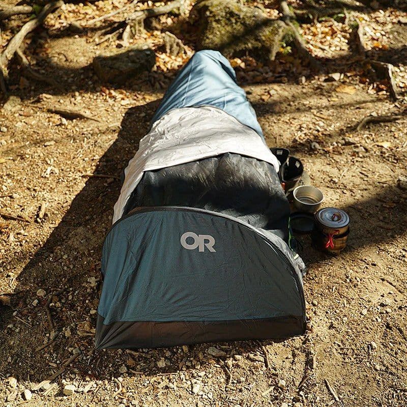 A bivy tent, sleeping bag, and cooking pots lie on a dirt path in a forest.