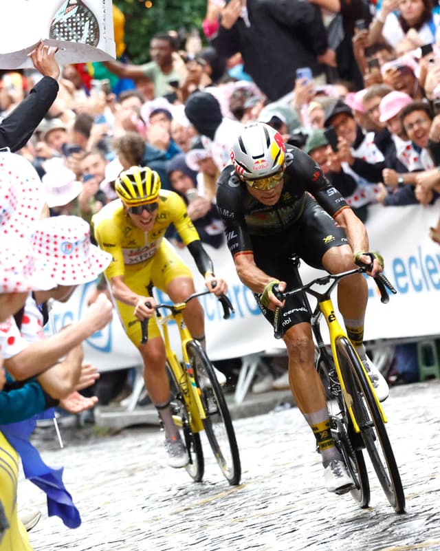 Two cyclists, one in a yellow jersey, race on a cobbled street past a cheering crowd.