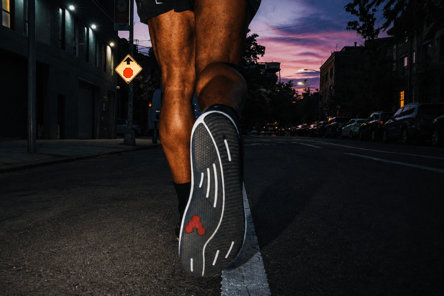 The sole of a running shoe and a person's leg mid-stride on an urban street at dusk, beneath a vibrant sky.
