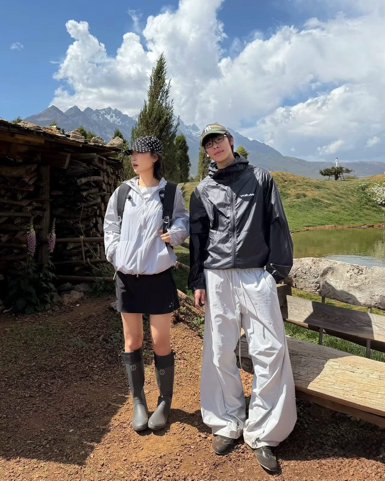 Two people in outdoor gear stand in a mountainous landscape with a lake and wooden structure.