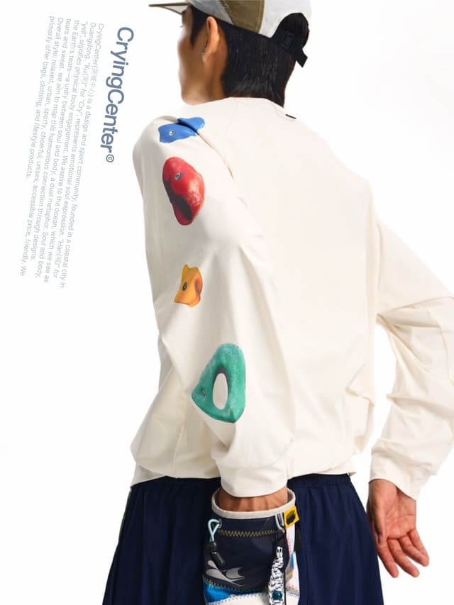 A person from behind wears a cream long-sleeved shirt with colorful rock climbing holds on the sleeves, a hat, and holds a small pouch. "CryingCenter" text is on the left.