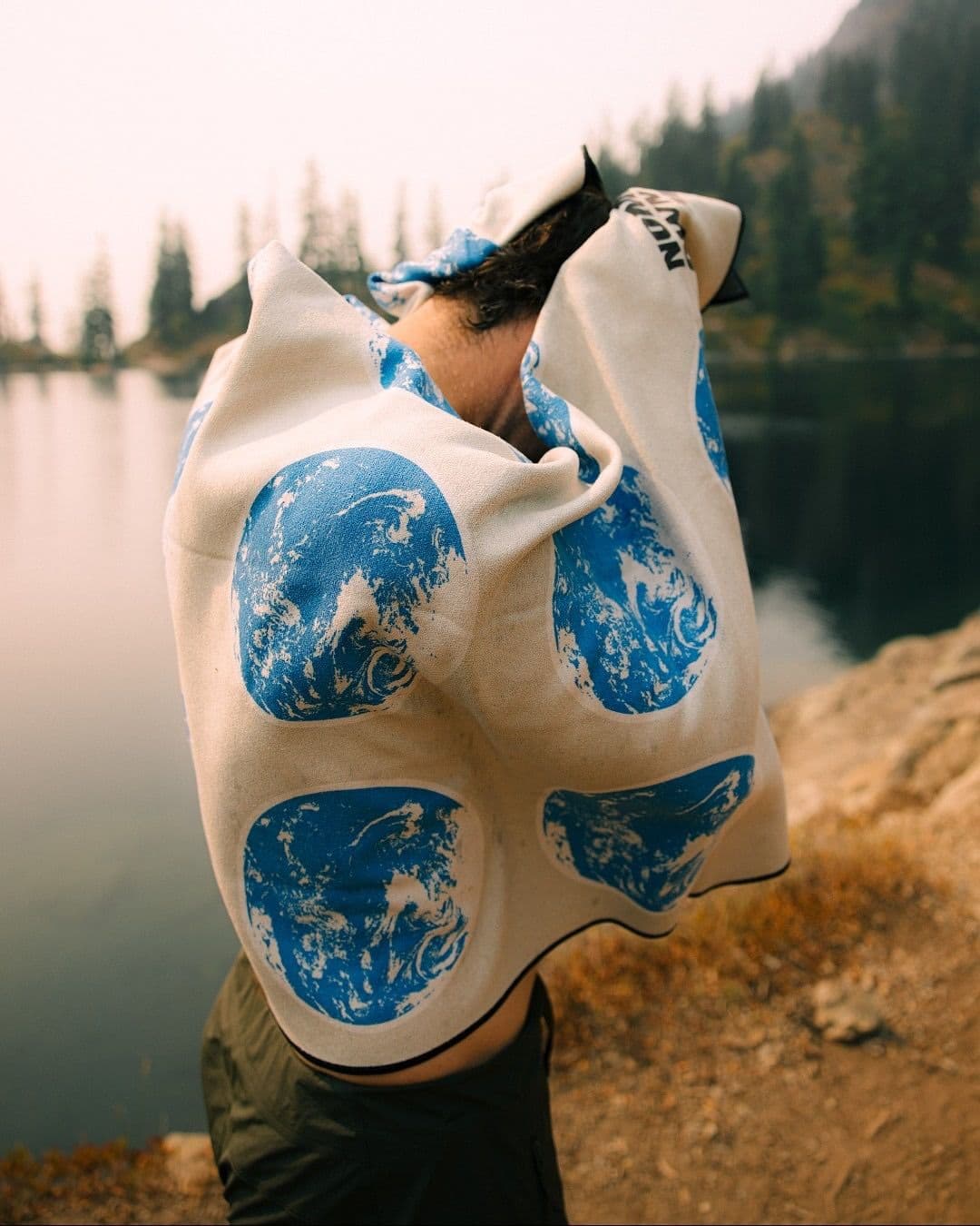 Person drying off with a cream towel patterned with blue earth globes, by a lake.