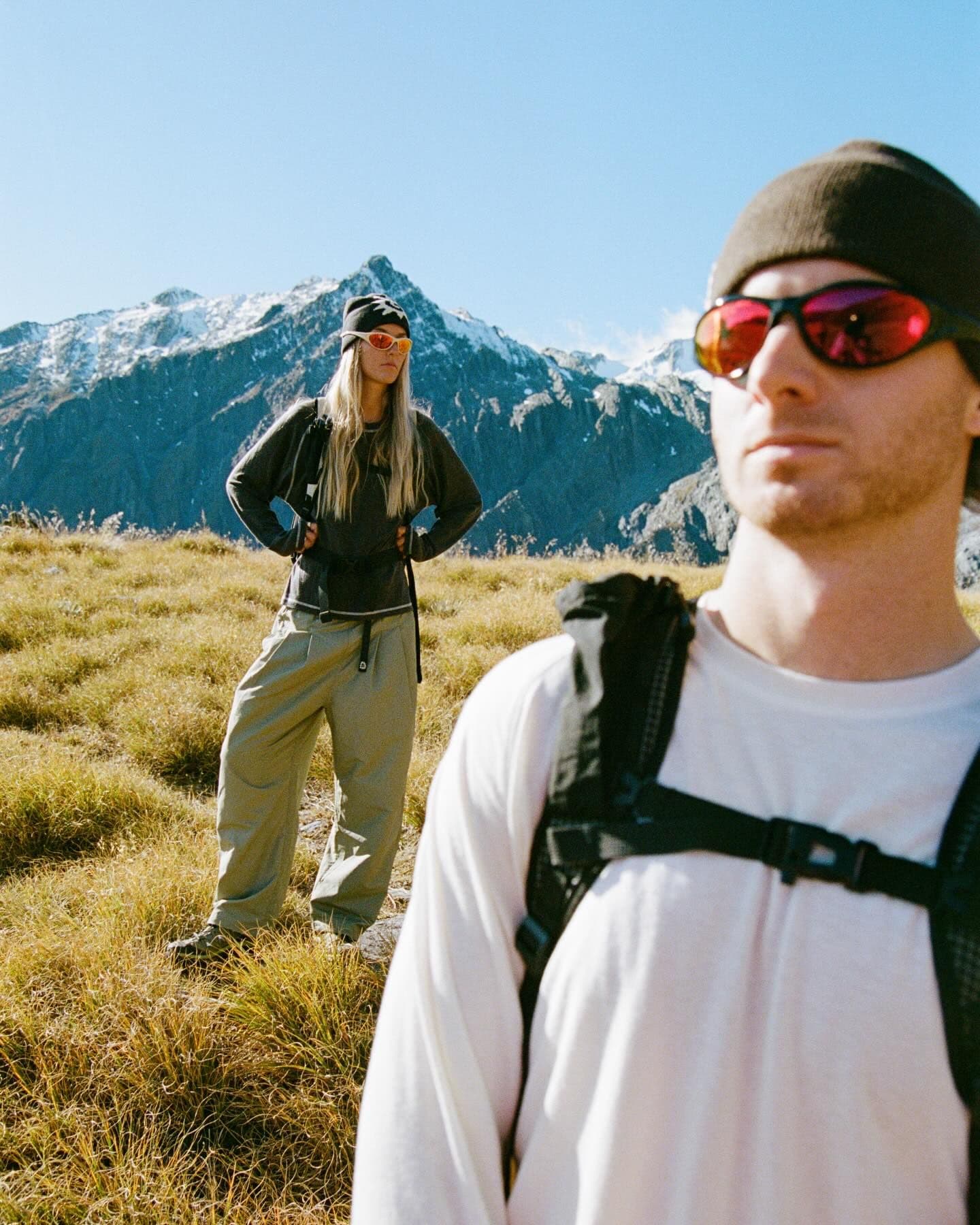 Two people in outdoor gear and reflective sunglasses stand in a sunny, golden field with snow-capped mountains under a clear blue sky.