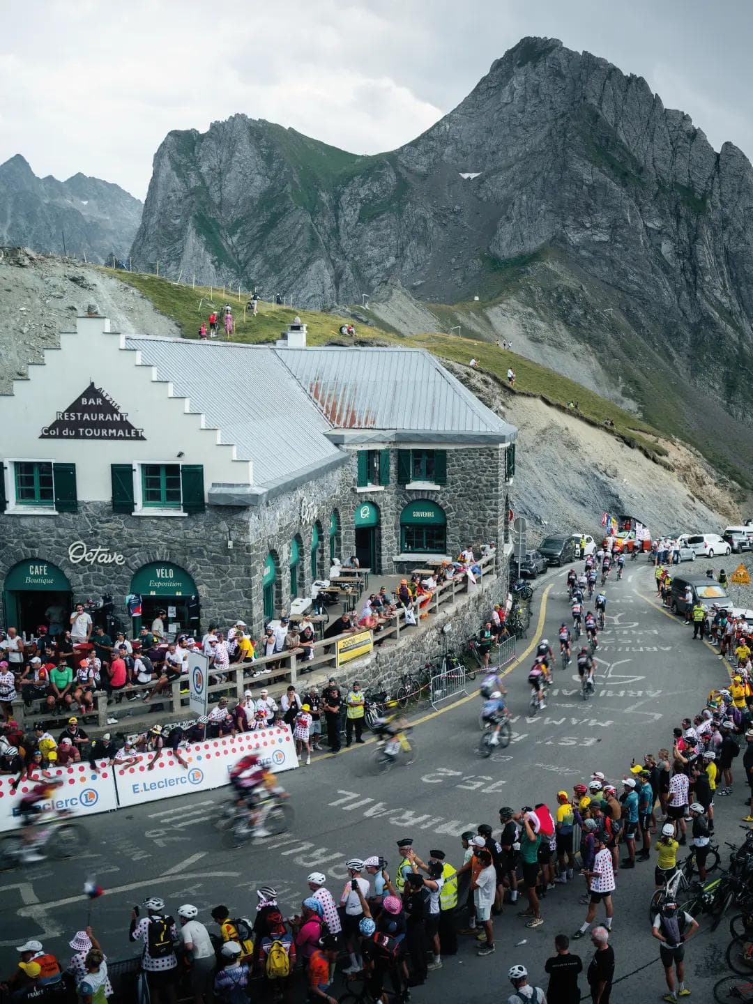 A bicycle race at Col du Tourmalet, with cyclists on a winding mountain road lined by many spectators and a stone building, all beneath rugged peaks.