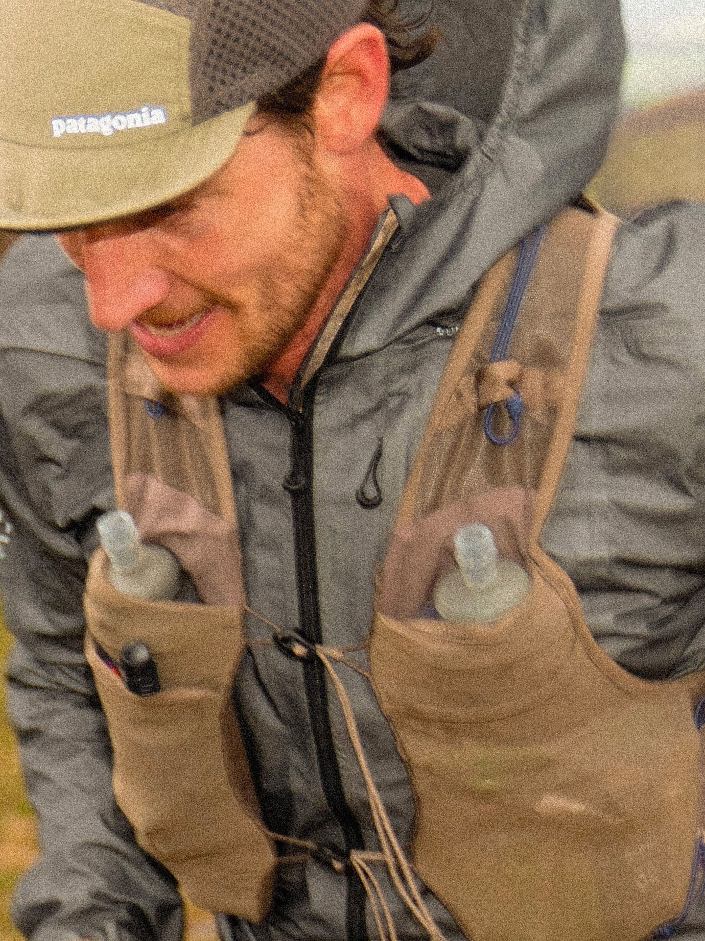 A man wearing a Patagonia cap, gray jacket, and a tan hydration vest with two water bottles.