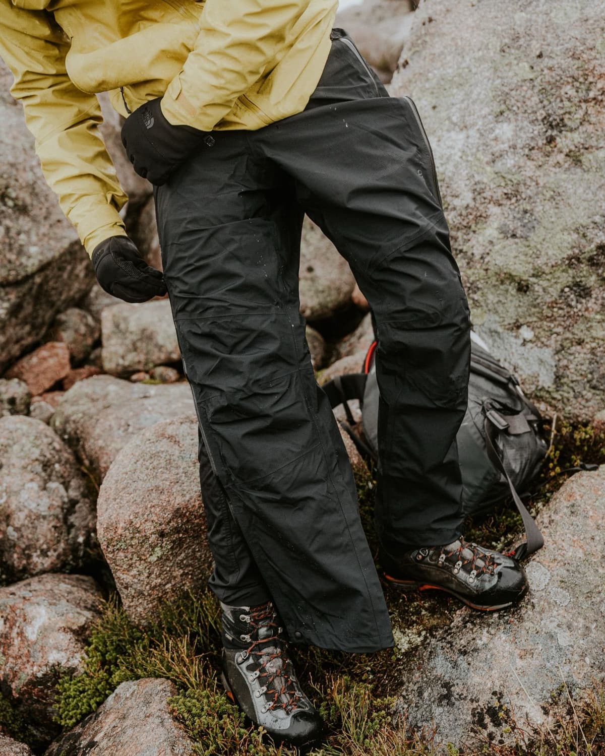 A person wearing a yellow jacket, black gloves, black waterproof pants, and hiking boots stands on rocky, mossy ground.