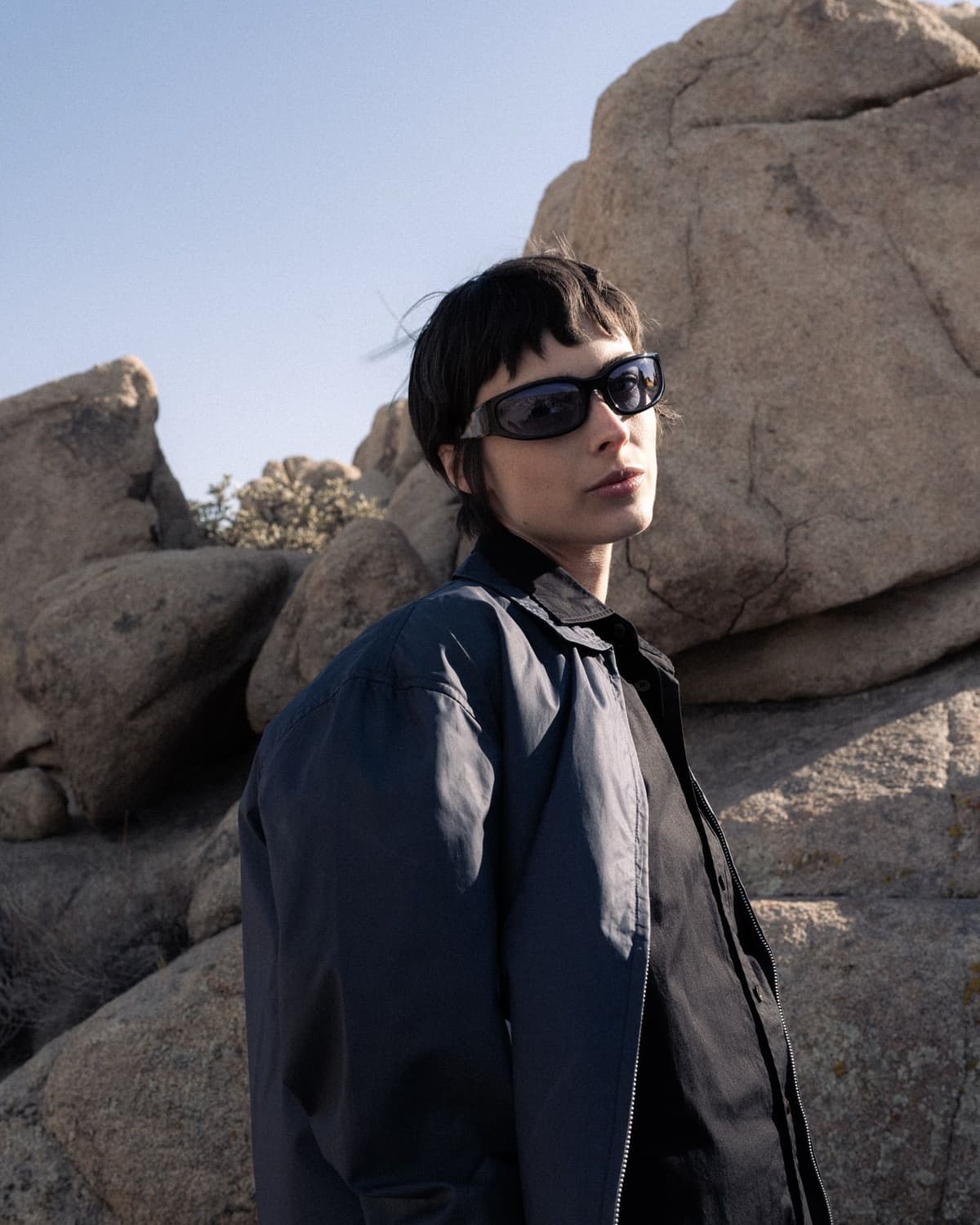 A person with short dark hair and sunglasses wearing a dark blue jacket stands in a rocky desert landscape.