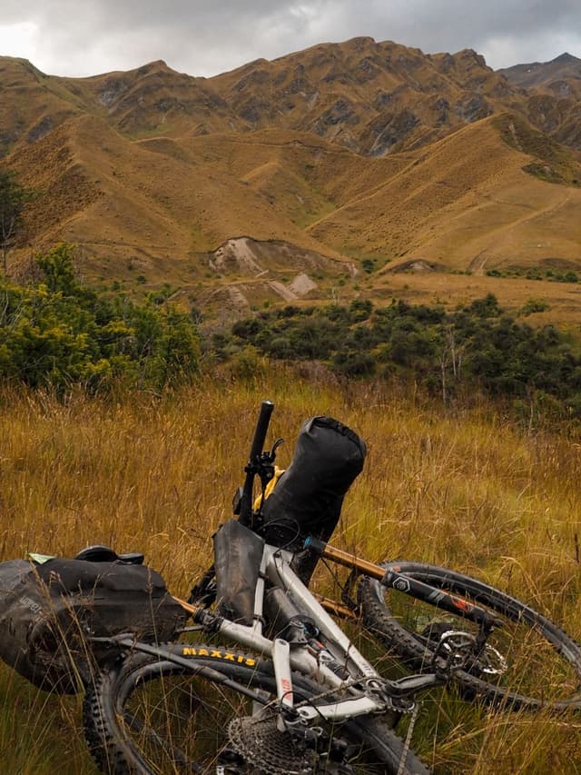 A bikepacking mountain bike rests in tall golden grass with rolling golden-brown mountains under a cloudy sky in the background.