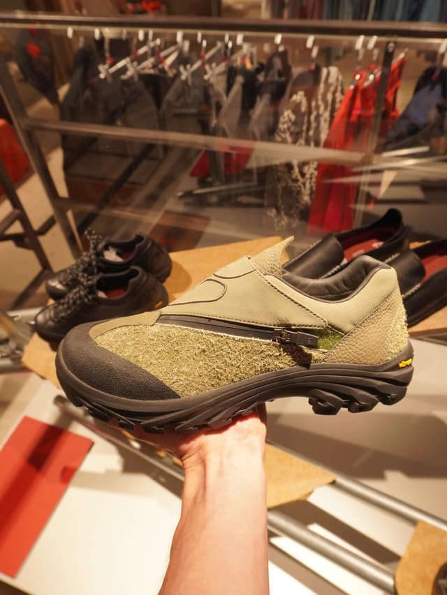 A hand holds up an olive green shoe with a zipper, textured sides, and a black Vibram sole.