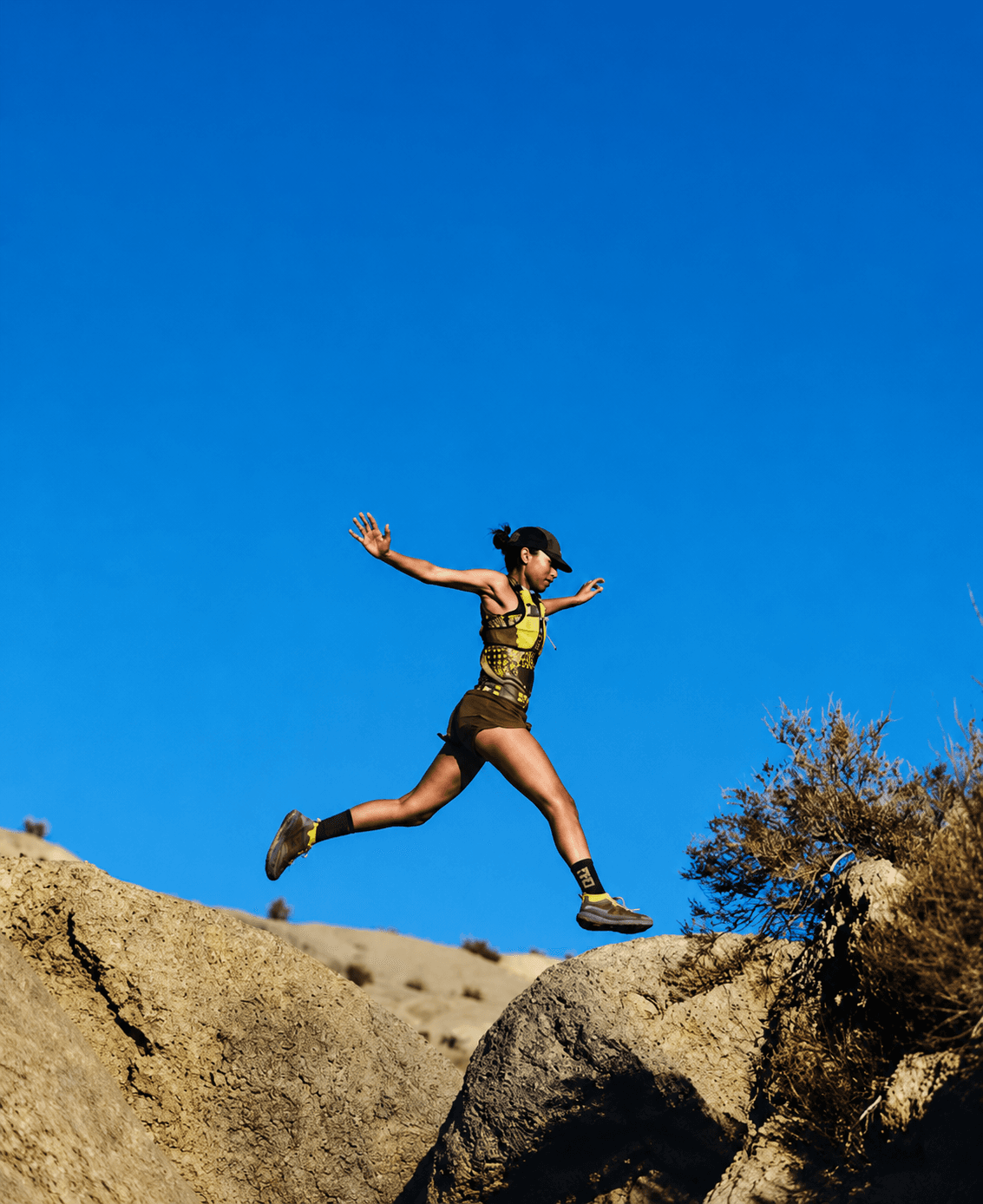 A woman trail runner leaps across rocks under a clear blue sky.